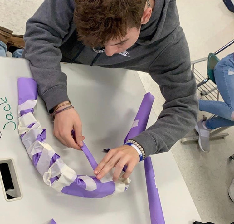 A student tapes a deer antler tine onto the main antler beam all made of purple construction paper and masking tape.