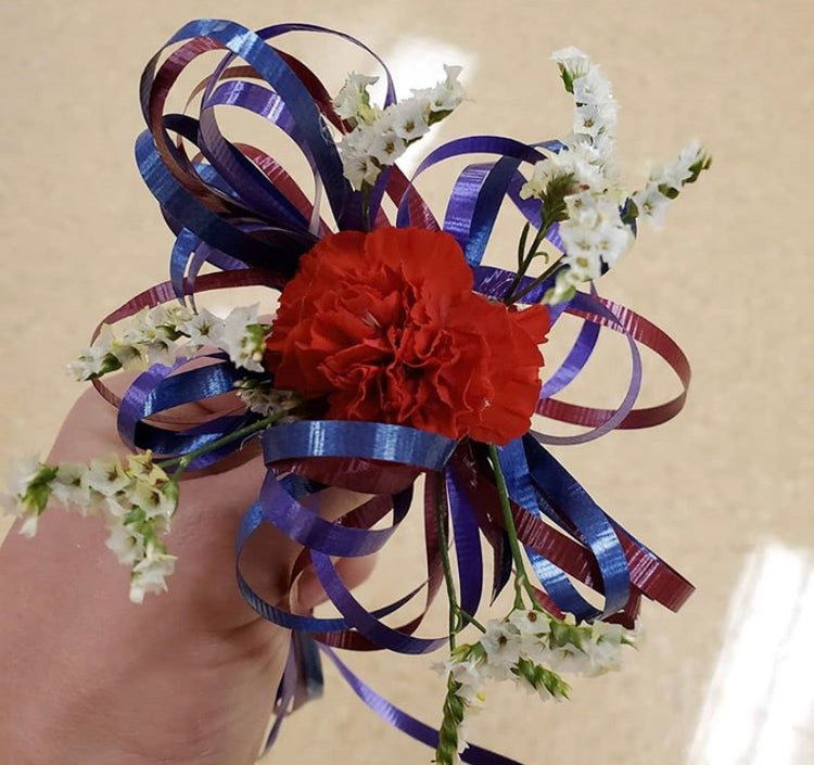 Student made corsage with red carnation, blue and burgundy ribbon loops, and small white flower accents