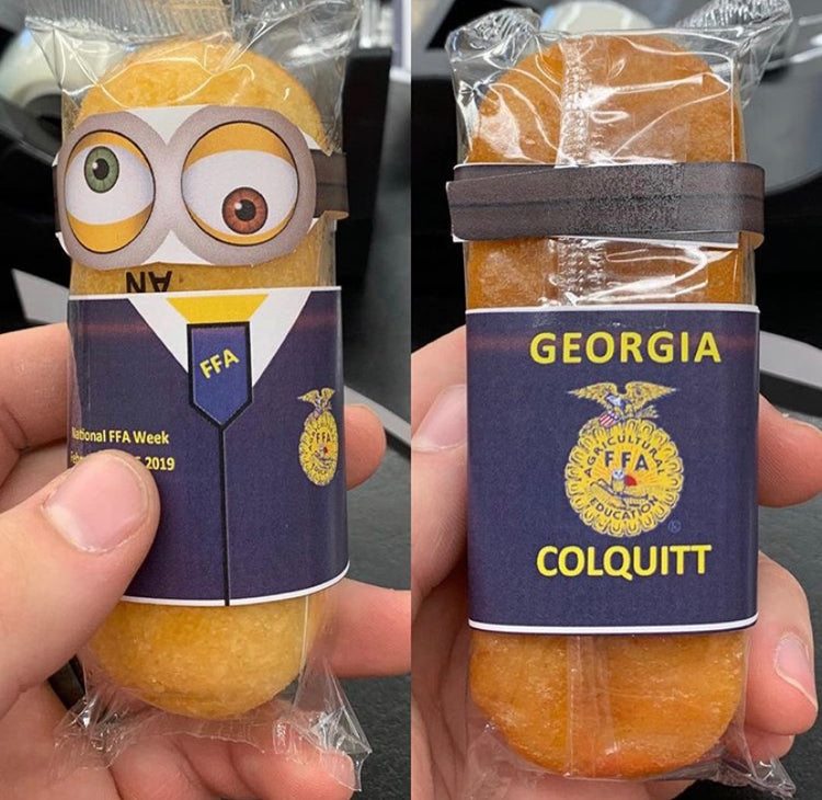 Yellow twinkie still wrapped in its clear wrapper has a paper cut out of an FFA jacket and goggled eyeballs taped to the exterior to look like a Minion in official dress.